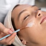 Mulher de olhos fechados recebendo a aplicação de um peeling facial em gel com pincel por uma esteticista.