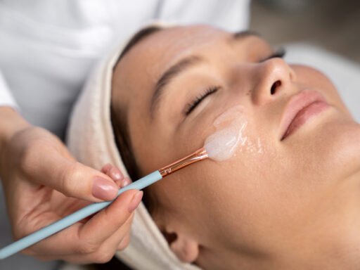 Mulher de olhos fechados recebendo a aplicação de um peeling facial em gel com pincel por uma esteticista.