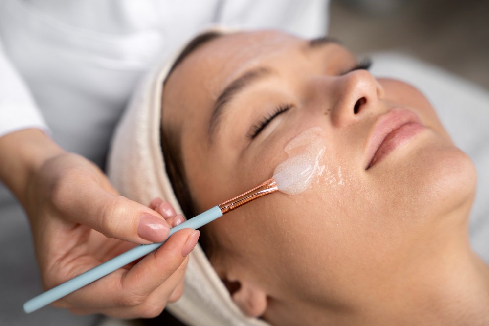 Mulher de olhos fechados recebendo a aplicação de um peeling facial em gel com pincel por uma esteticista.